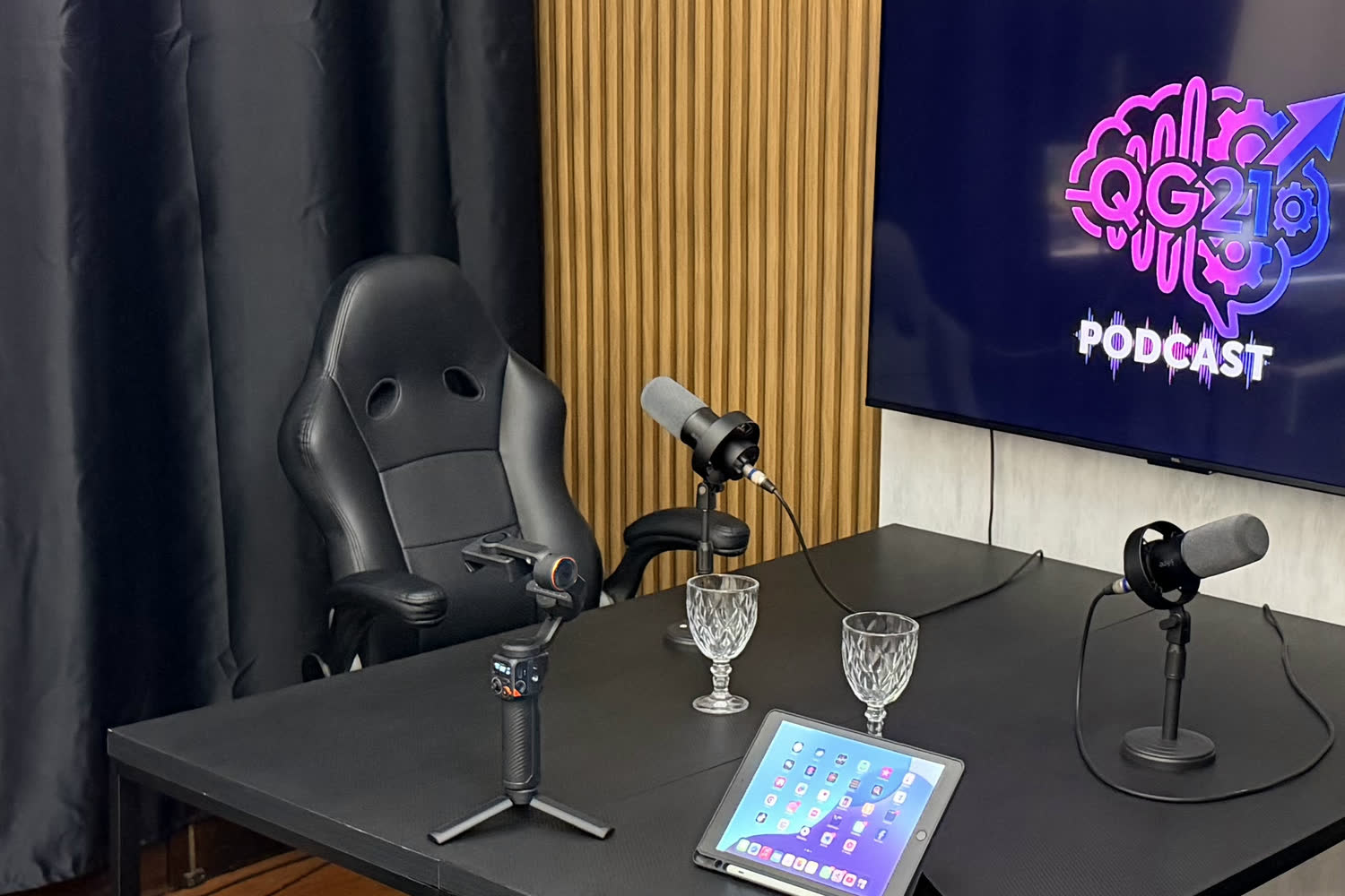 Mesa de gravação do estúdio de podcast da QG21 Studio com microfones, cadeiras e a TV com o logo do programa