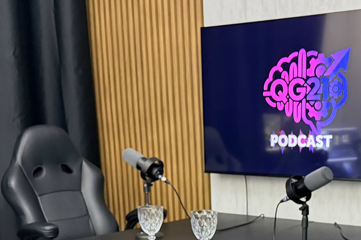 Bancada do estúdio de podcast da QG21 Studio com dois microfones e o logo QG21 Podcast na TV